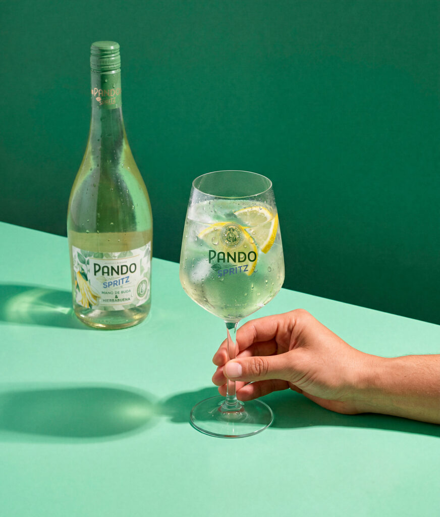 Mano sujetando una copa de Pando Spritz junto a la botella